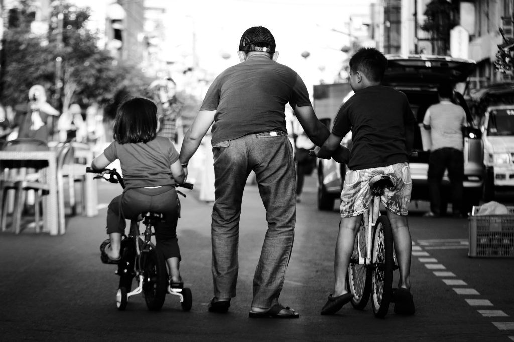 A father guides his children on bicycles down an urban street in black and white.